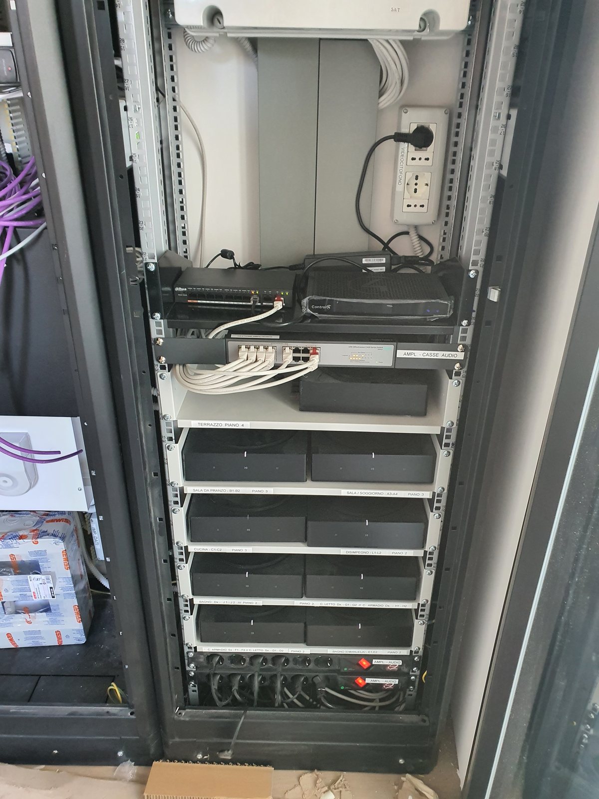 Armadio rack con controller domotico, switch di rete e amplificatori audio etichettati per ambiente